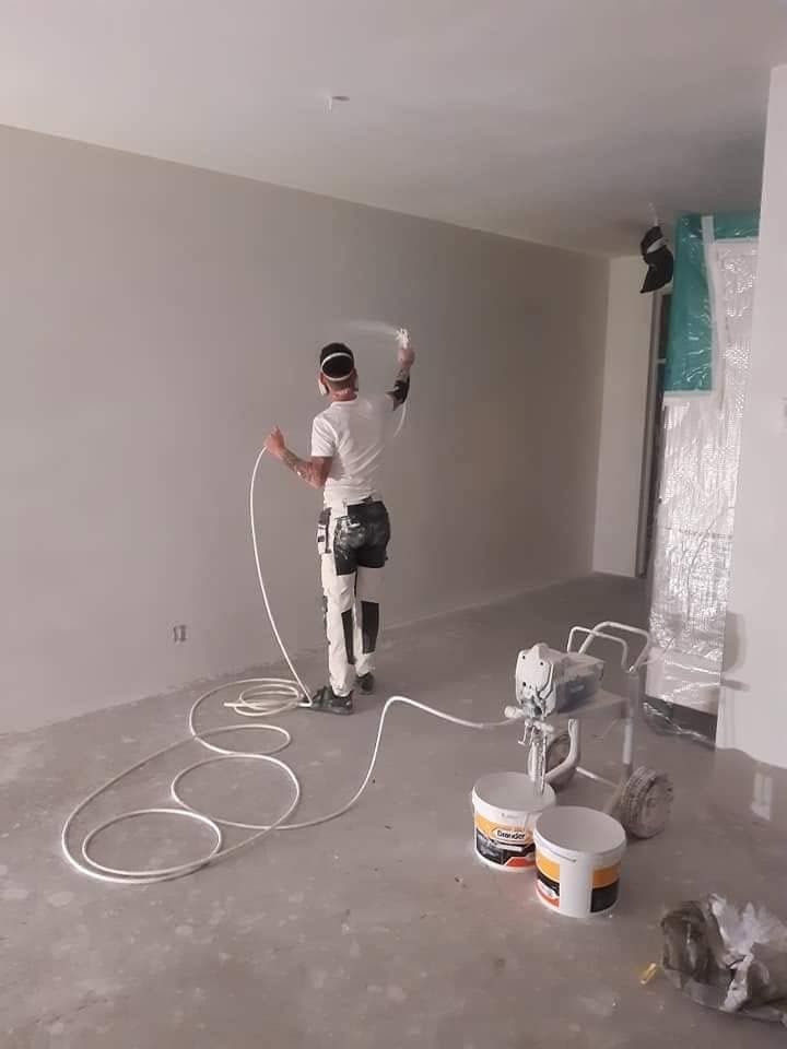 Tijdens het werk: schilder spuit de wanden met een airless spuit; de emmers staan op de vloer. (Facebook, 10 januari 2021)