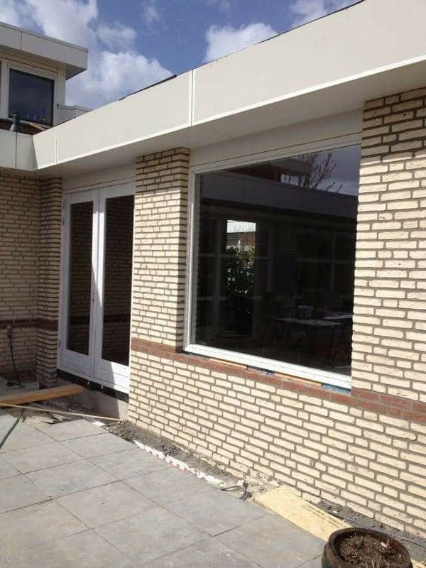 Aanbouw met plat dak: wit boeiboord, openslaande deuren en een groot raam in lichte baksteen. (jan 2021)