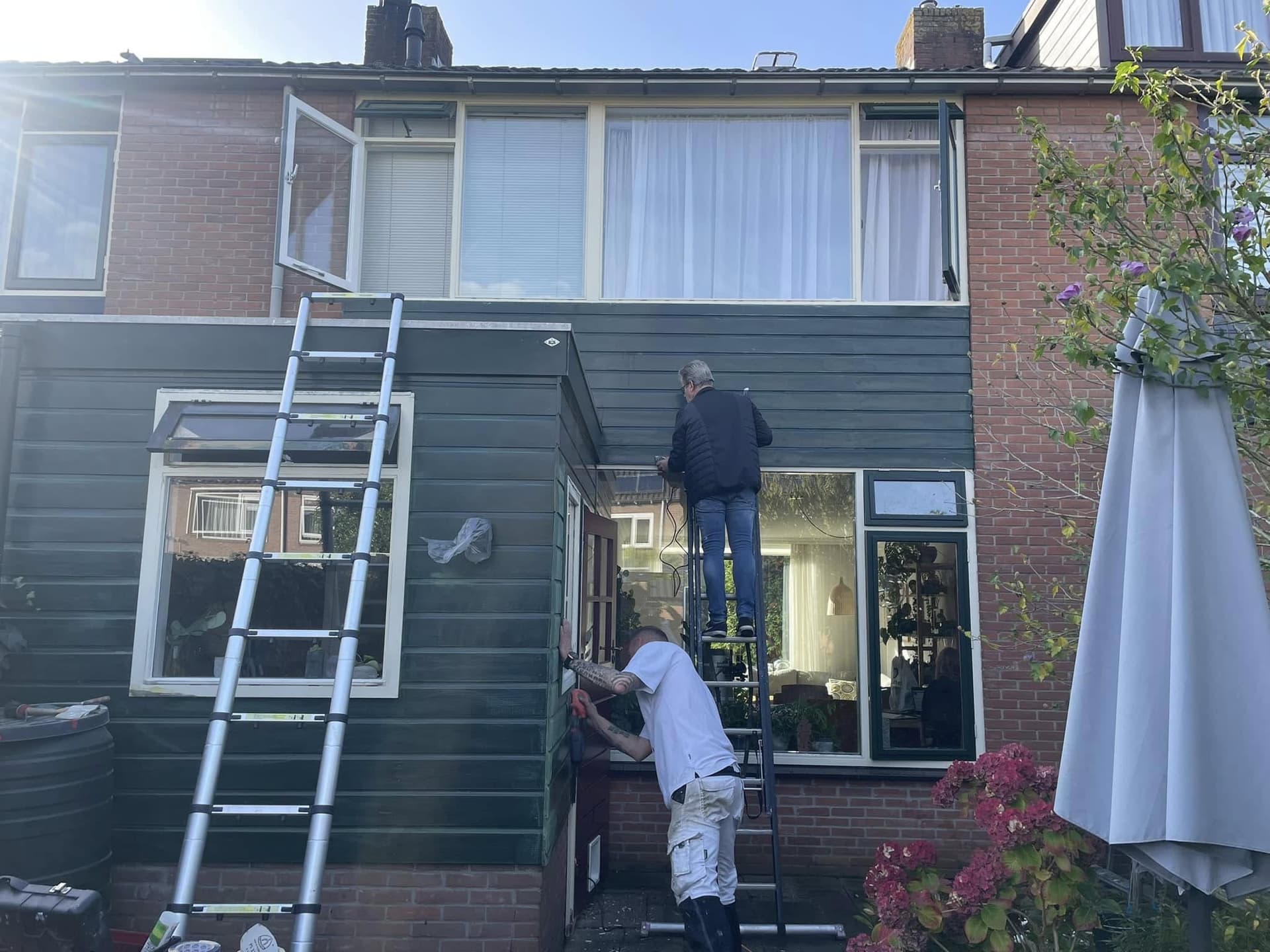 Tijdens het werk: twee schilders op ladders aan de achtergevel met de houten aanbouw. (Facebook, 16 oktober 2024)